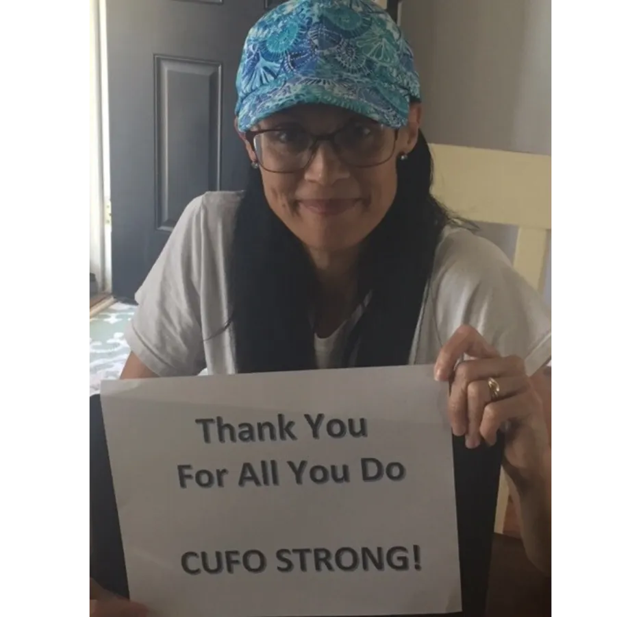 A woman wearing a blue paisley baseball cap holds a sign that says, "Thank you for all you do. CUFO strong!"