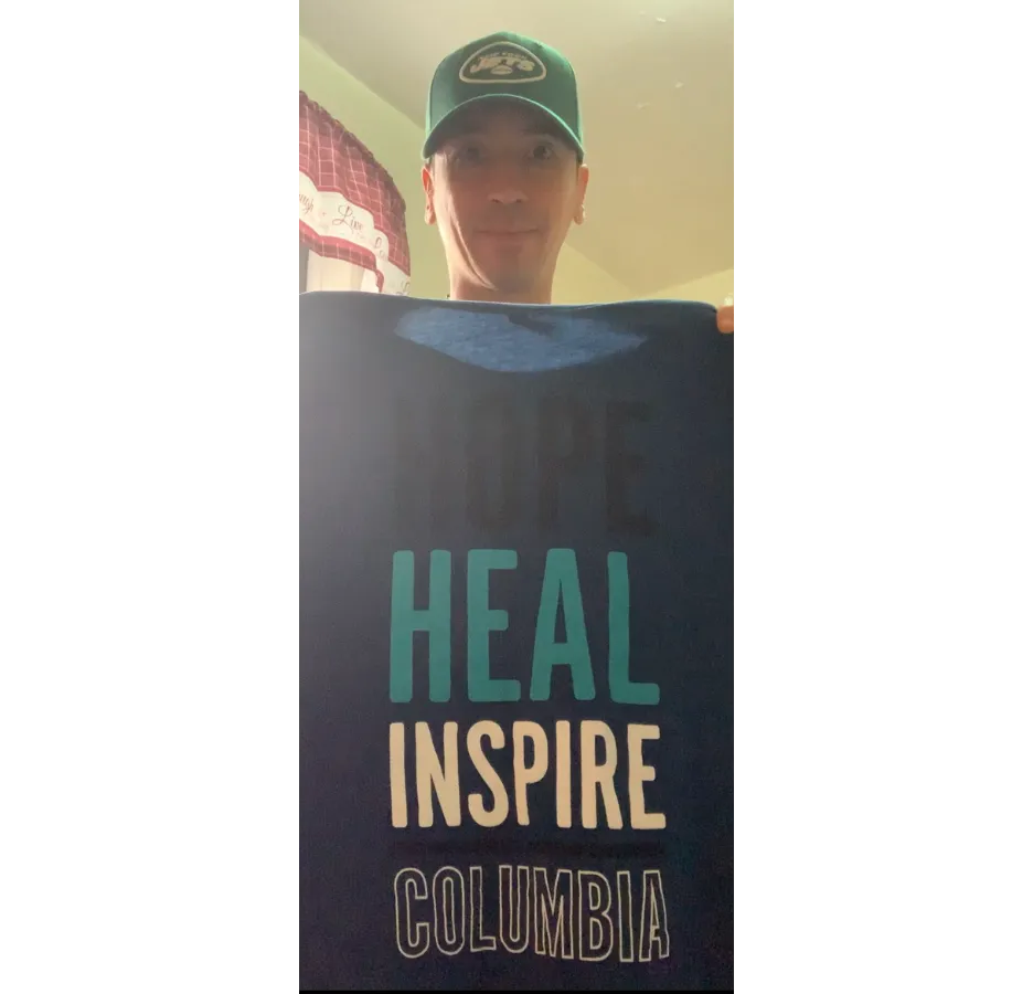 A man wearing a green baseball cap holds a shirt that says, "Hope, heal, inspire, Columbia"
