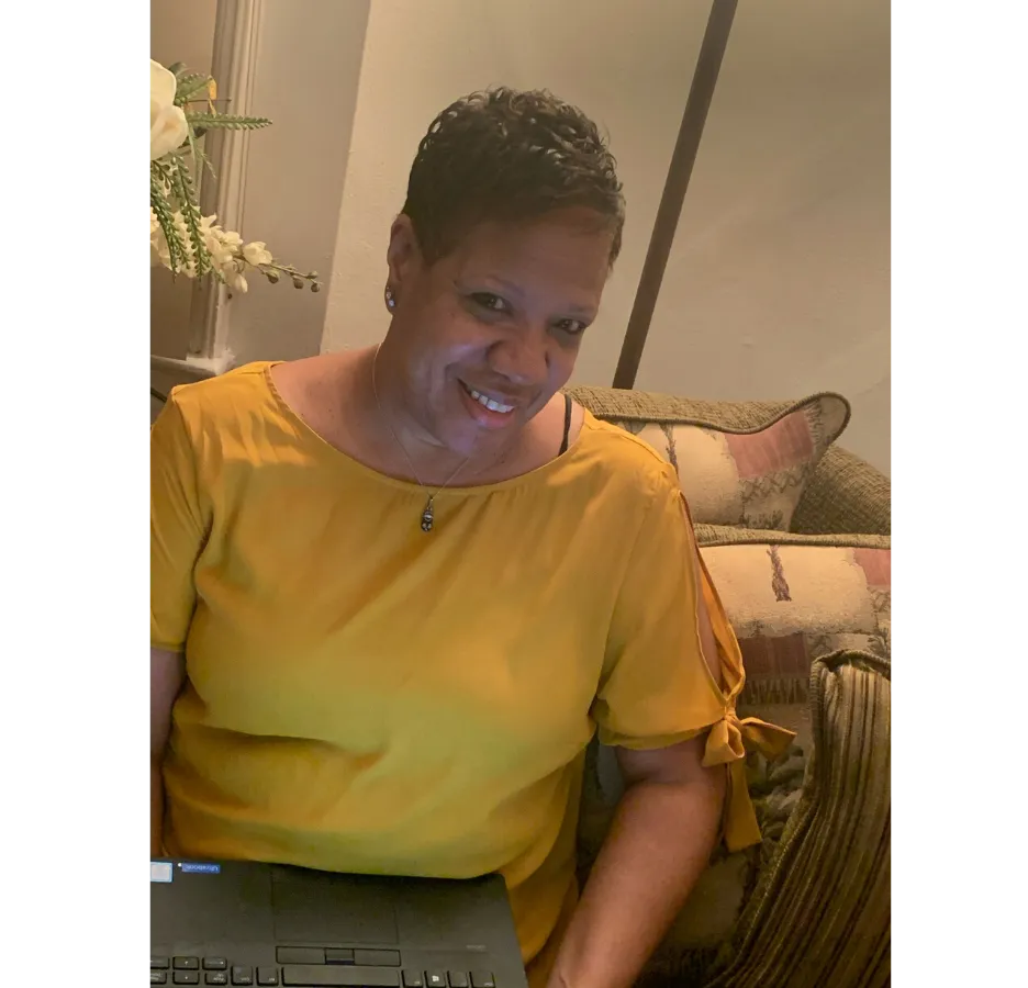 A woman wearing a yellow shirt with a laptop on her lap smiles at the camera