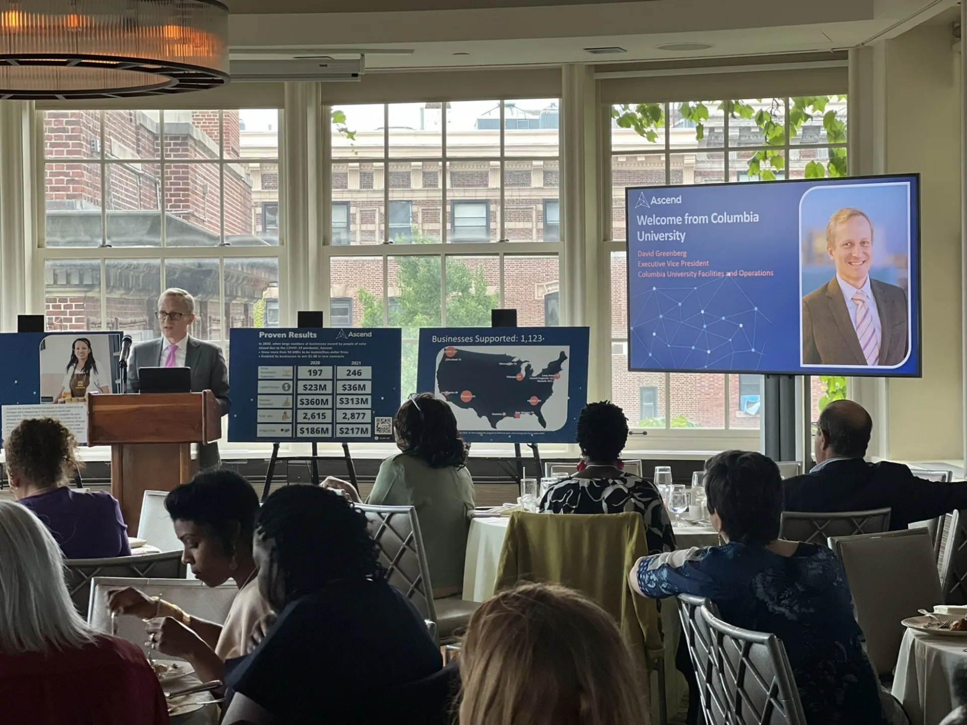 People sitting at tables in a room with white walls and big windows, listening to a speaker at a podium, with a digital monitor that has a photo of speaker and text that says "Welcome from Columbia University"