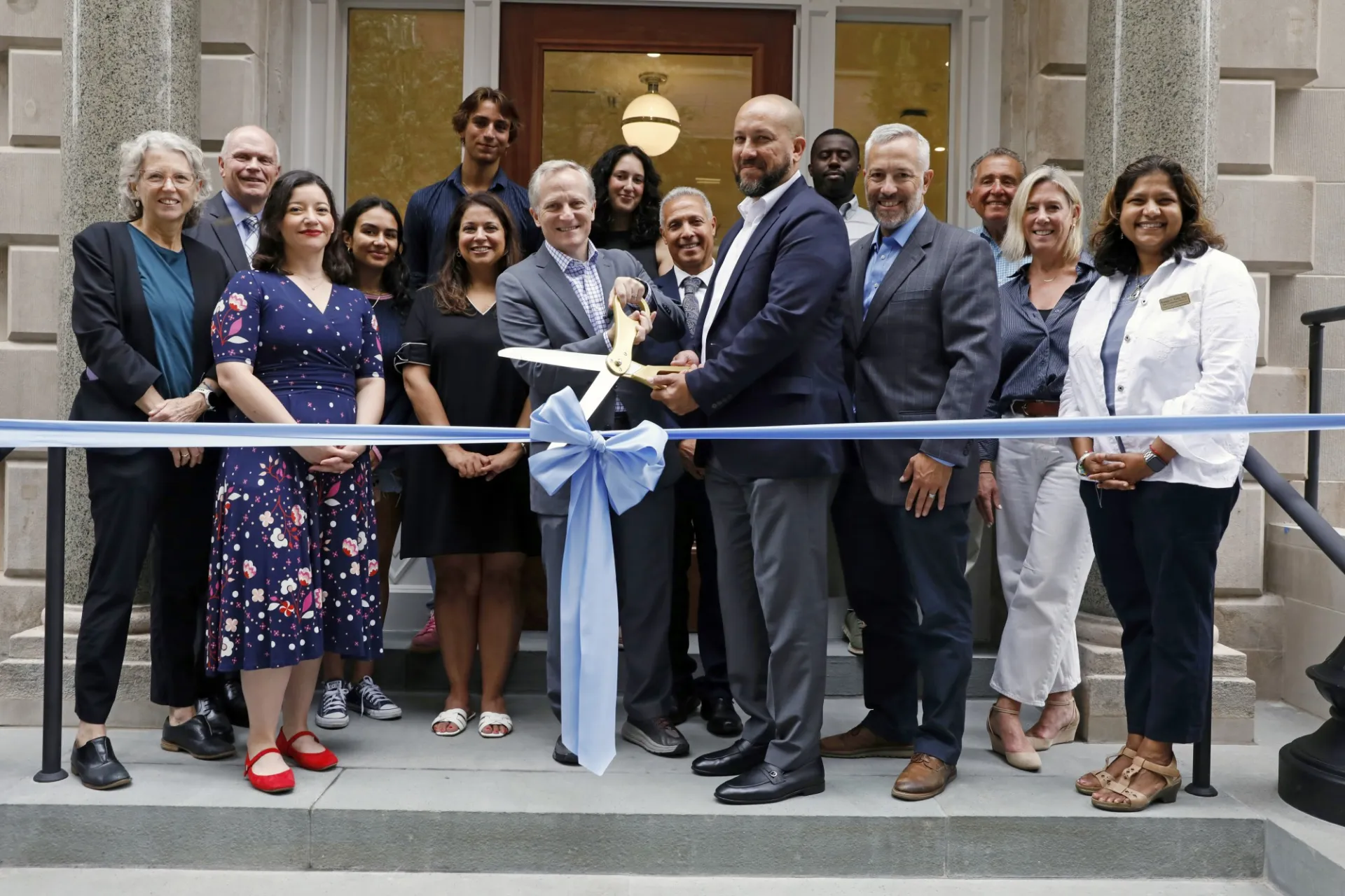 Columbia University Leaders gather in front of the doorstep of 611 W. 112th Street and are about to cut a large, blue ribbon with large, gold scissors.