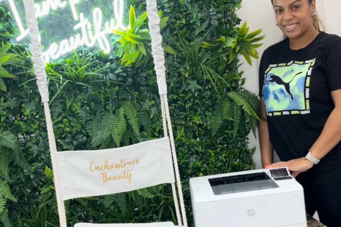 A person stands next to a new HP printer and a white swing that says Enchantress Beauty that is in front of a foliage wall.