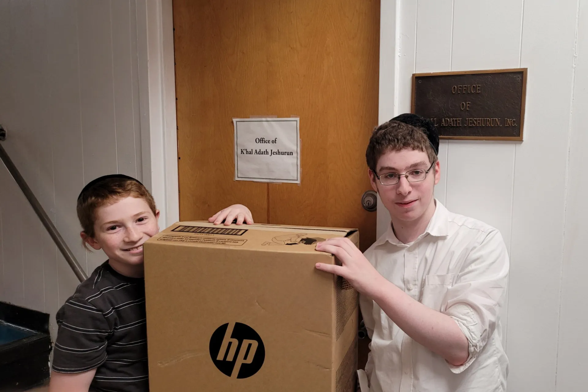Two people hold up a HP printer that is in its box in the hallway outside of their office.