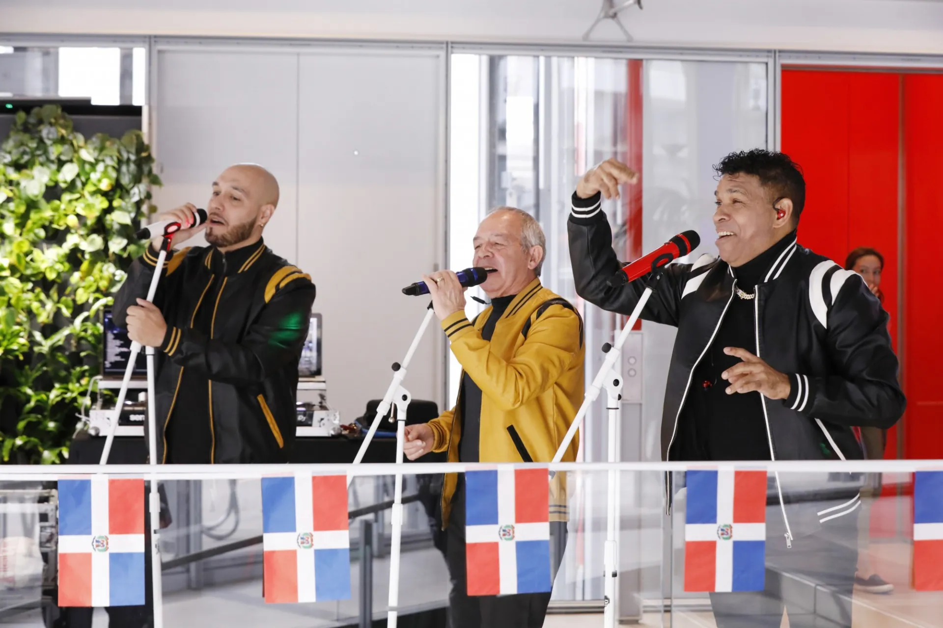 Three people sing into microphones. Small Dominican flags are in the foreground of the photo.