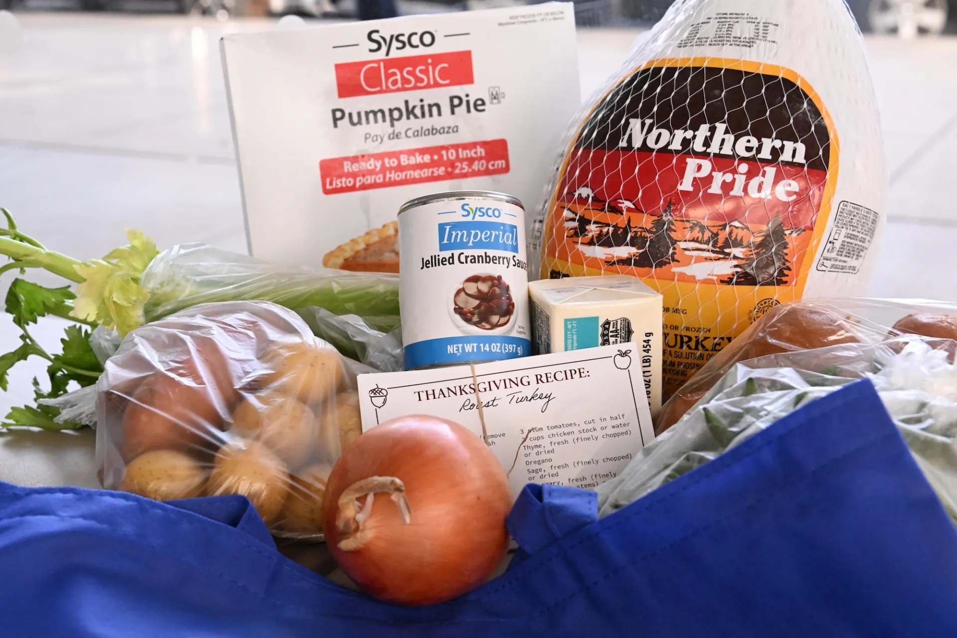 Thanksgiving food items including a frozen turkey, pumpkin pie box, cranberry sauce can, butter, fresh vegetables, and a recipe card.