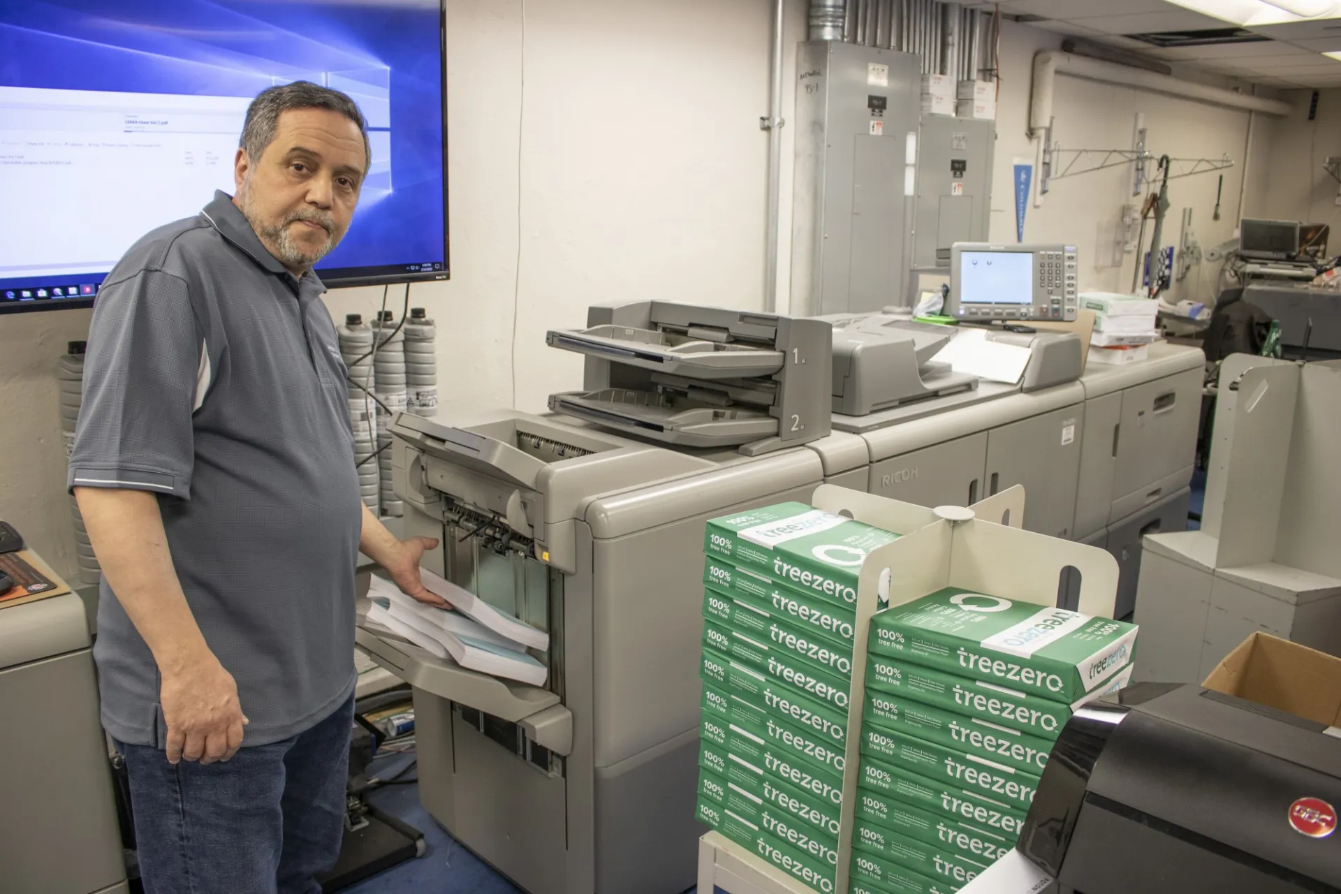 A man in a grey polo stands at the end of a large printing and copying device.
