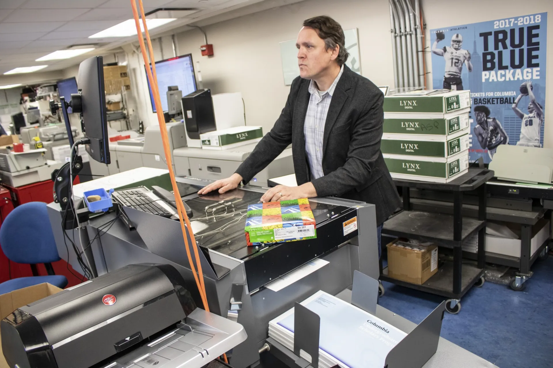 A man in a sport jacket stands at a desk facing a computer monitor.