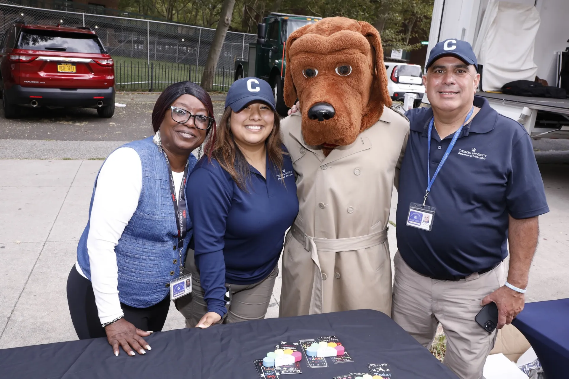 Three people stand behind a table smiling. Someone in a dog in trench coat mascot costume stands with them.
