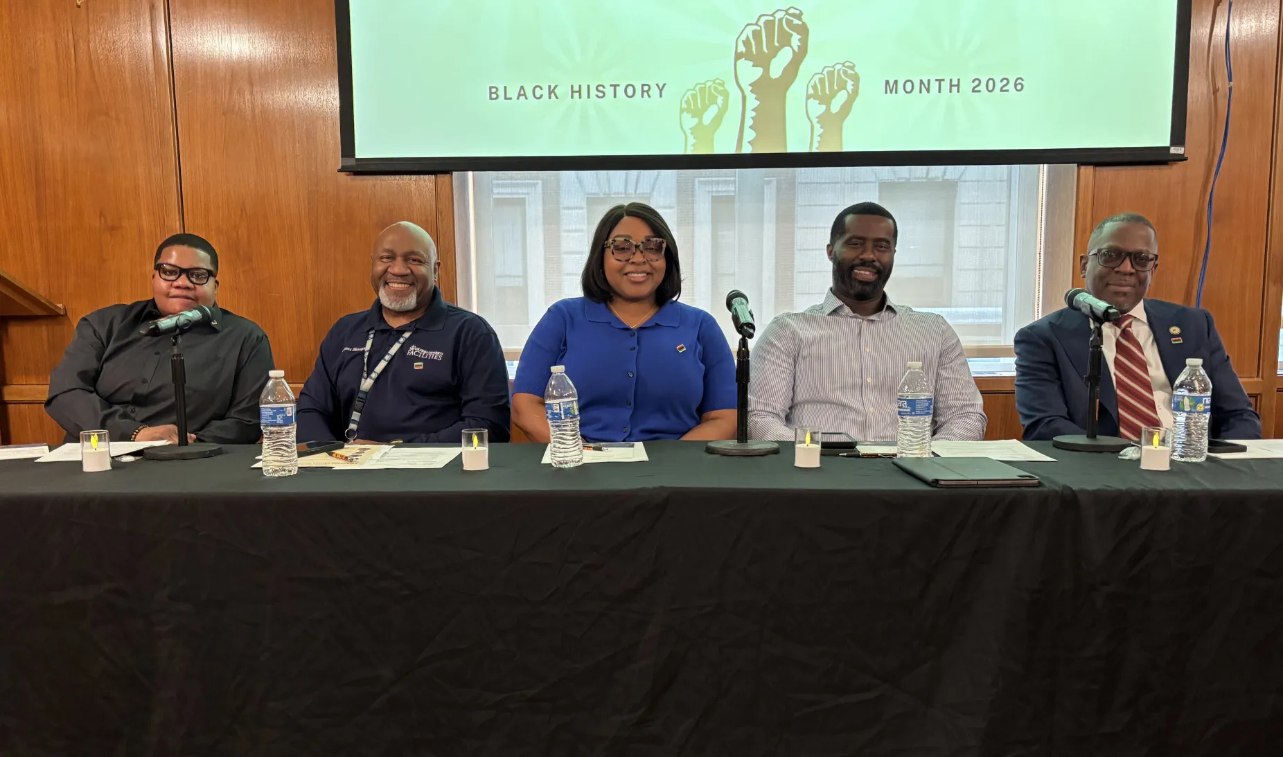 Five people sitting a table with three microphones. Behind is a PowerPoint Projection that says, "Black History Month 2026"