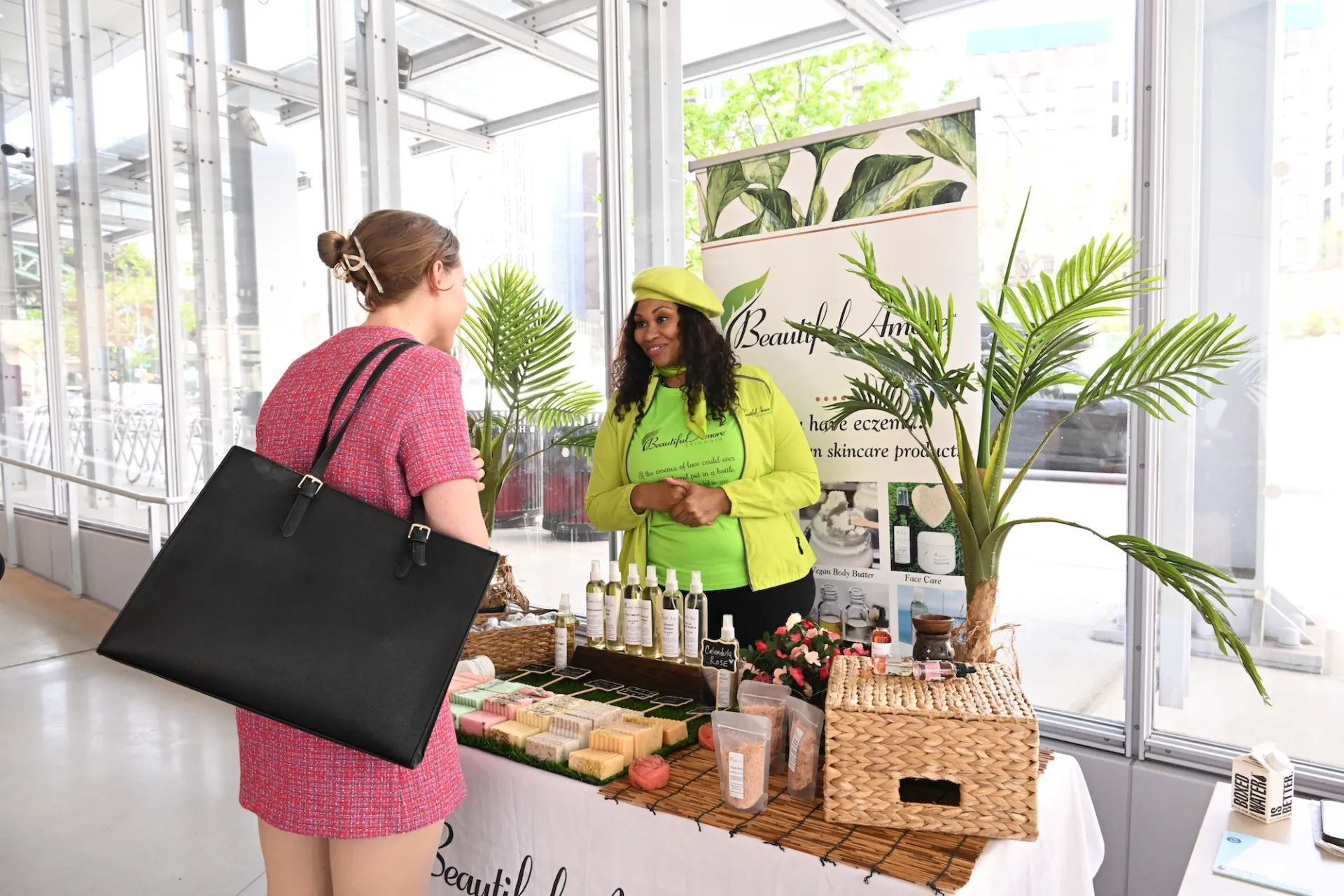 A woman in a pink dress with a large black handbag stands at a booth speaking with another woman wearing a bright green shirt and hat. The booth displays skincare products, including bottles and bars of soap, along with wicker baskets and decorative plants. A banner behind the booth reads 'Beautiful Amore Skincare' and mentions products for eczema and other skin conditions. Large windows in the background reveal trees and buildings outside.