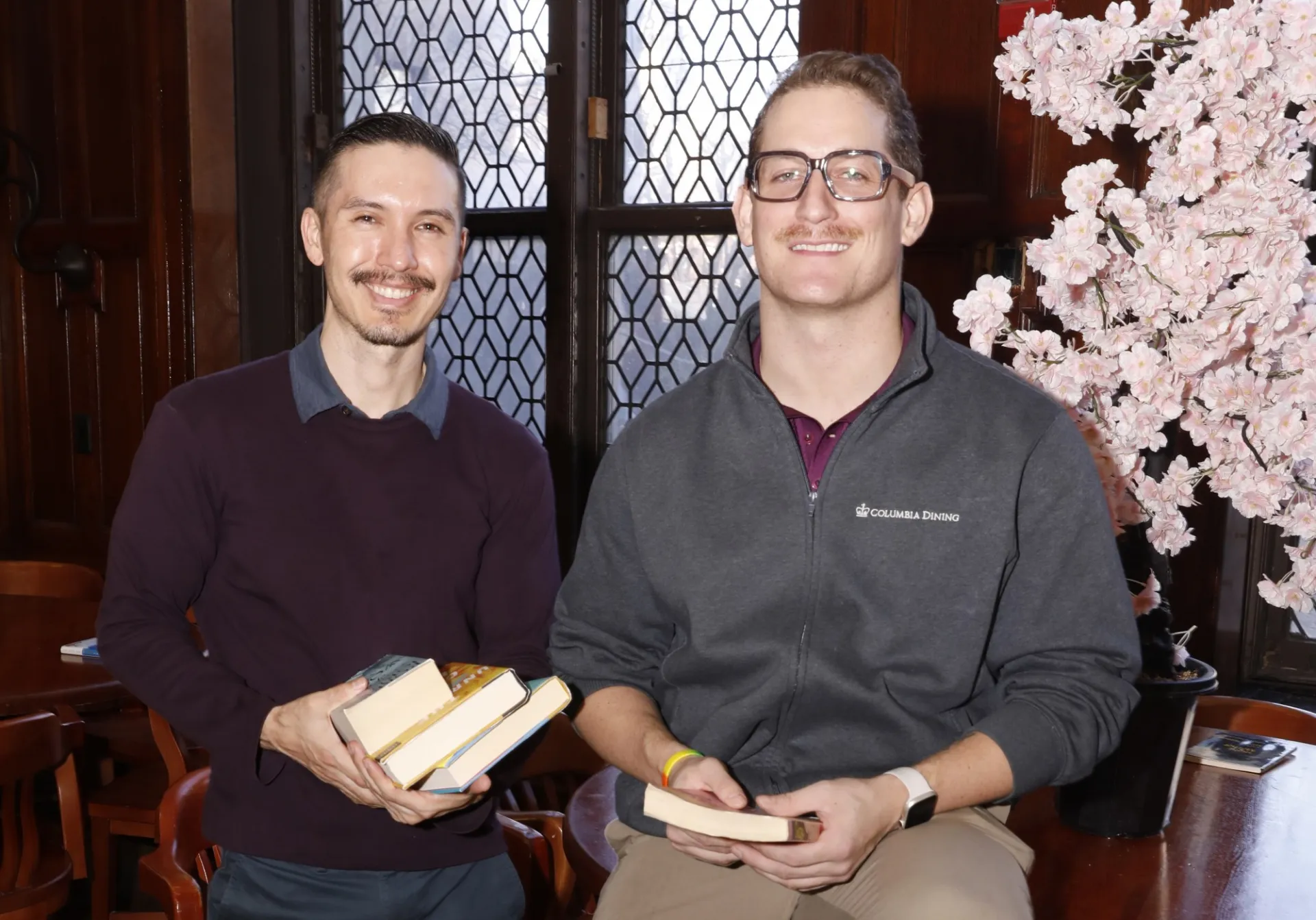 Two people sit indoors by a window; one holds books, the other wears a Columbia Business School sweatshirt and reads. A pink flowering tree is nearby.