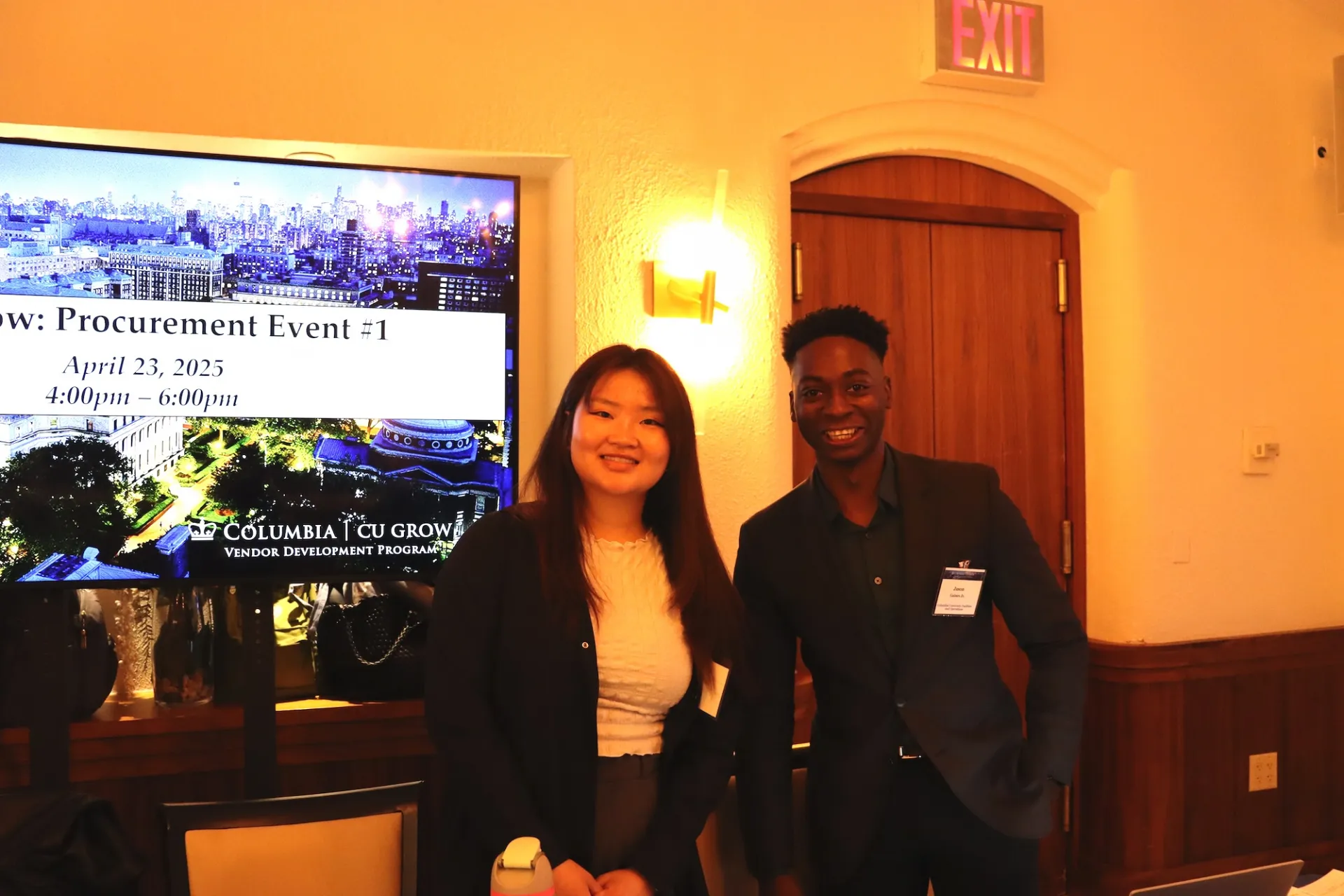 Two individuals stand in a room with a wooden door and an illuminated exit sign above it. Behind them, a large screen displays event information: 'Procurement Event #1, April 23, 2025, 4:00pm - 6:00pm' along with the Columbia CU Grow Vendor Development Program logo. The screen background features a cityscape image.