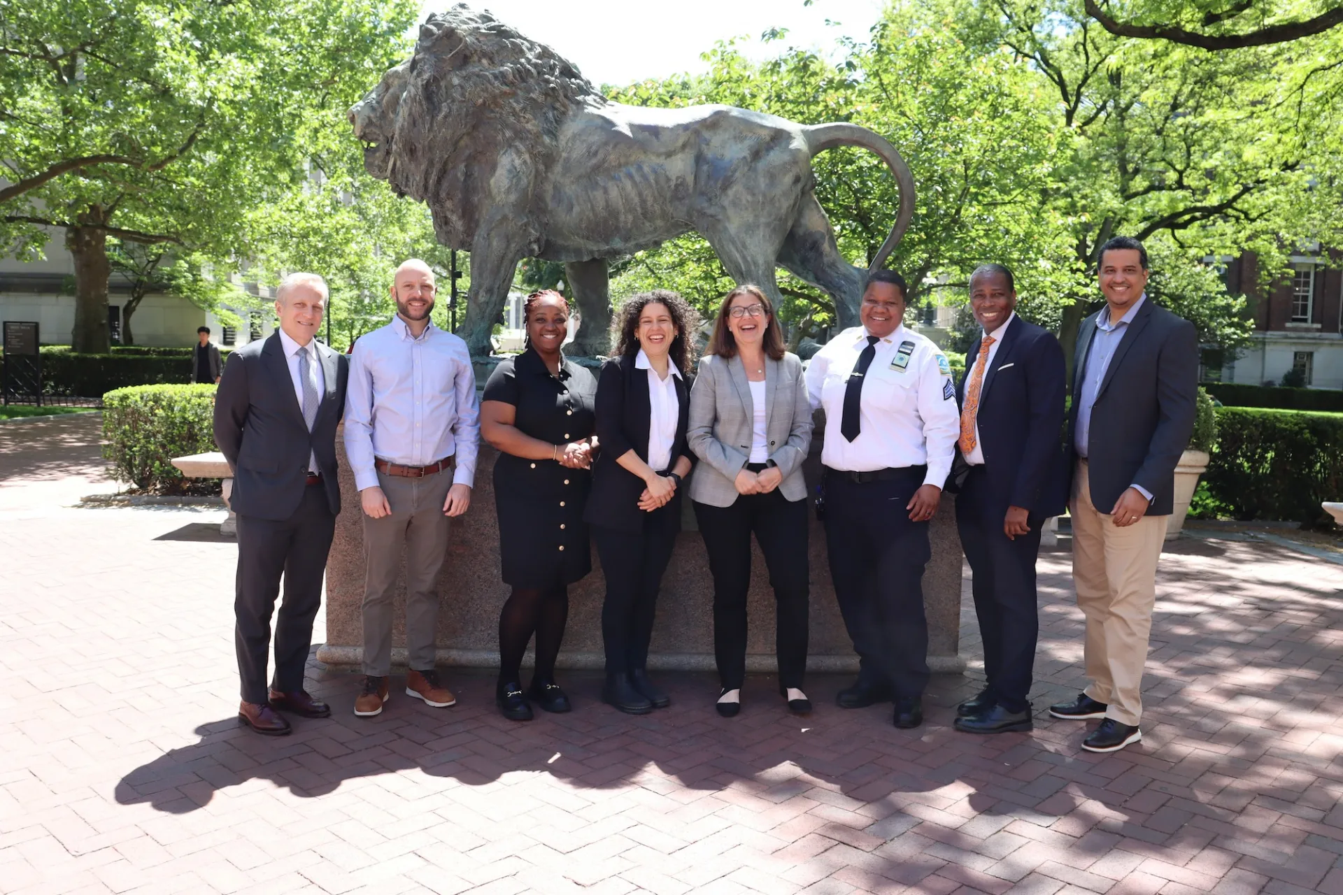 Eight people stand smiling in front of Scholar Lion on Columbia's Morningside Campus