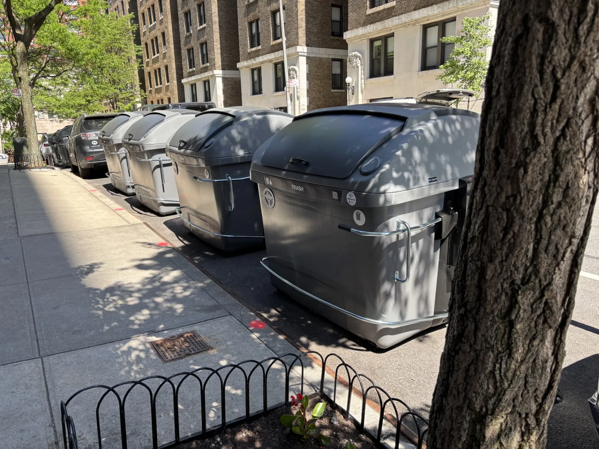 A row of large, gray garbage bins lined up along a city sidewalk next to a residential building. The bins are modern and have closed lids. There is a tree in the foreground on the right side, casting shadows on the sidewalk. The street and buildings are visible in the background.