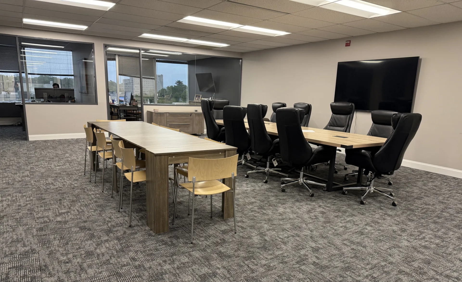 Modern office meeting space with two wooden tables: one long rectangular table with light wooden chairs and another set of tables arranged for a conference with black high-back swivel chairs. A large flat-screen monitor is mounted on the wall, and windows in the background provide natural light.