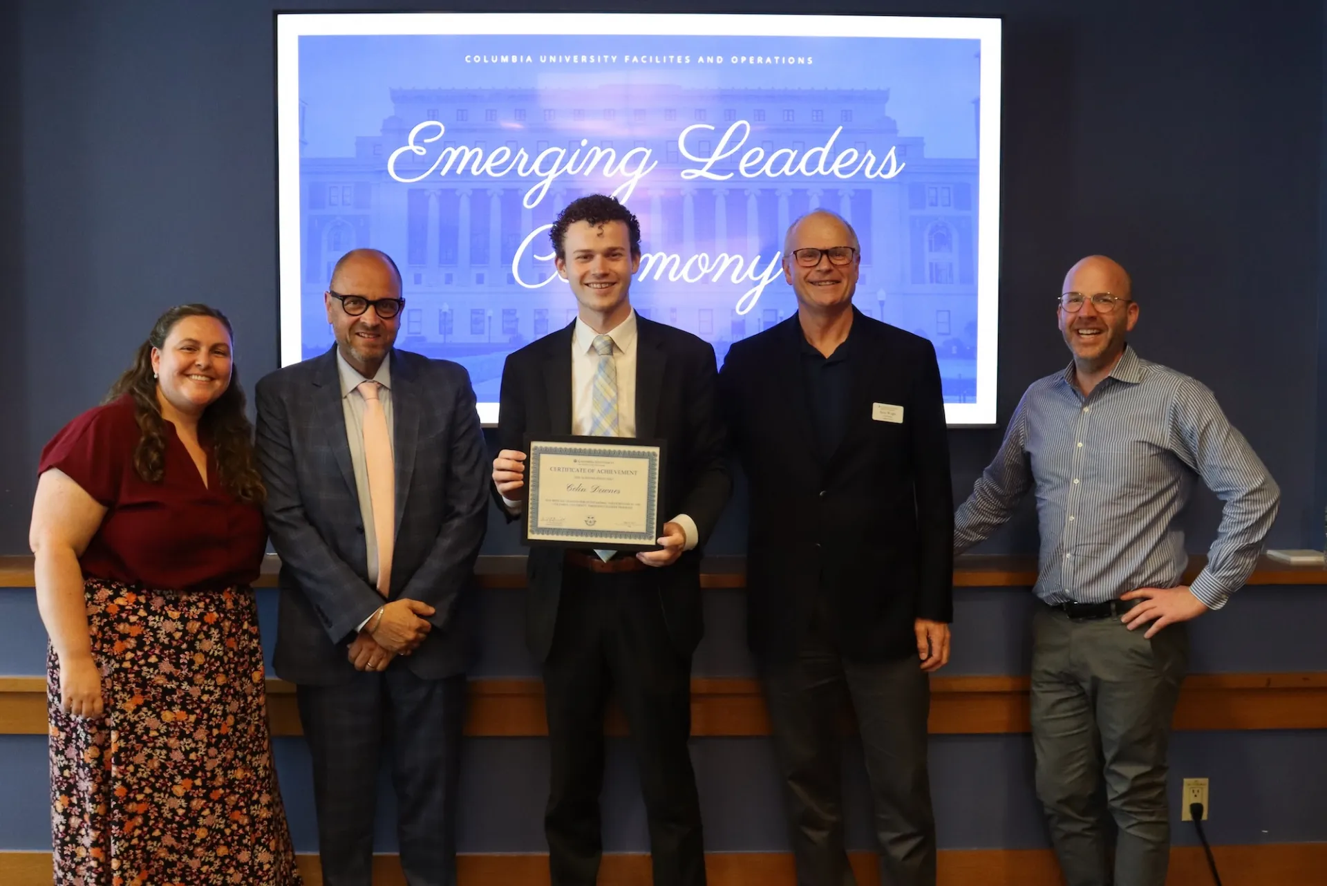 Five people stand in front of a screen reading 'Emerging Leaders Ceremony' at Columbia University; the center person holds a framed certificate.