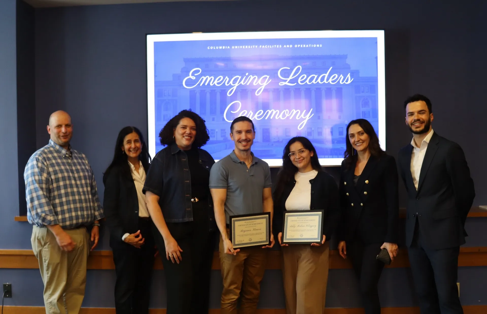 Seven people stand in front of a screen reading 'Emerging Leaders Ceremony' at Columbia University Facilities and Operations; two in the center hold framed certificates.