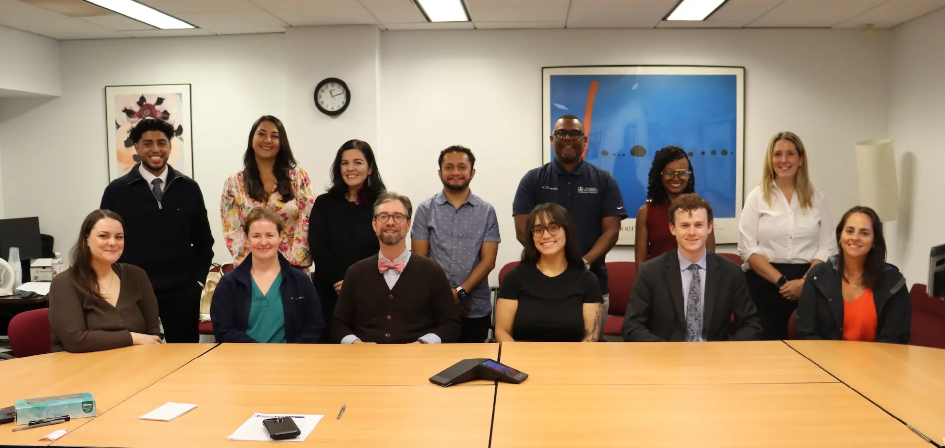 13 people (6 sitting and 7 standing behind them) smile and pose for a photo. Conference room table in the foreground and art and a clock behind them.