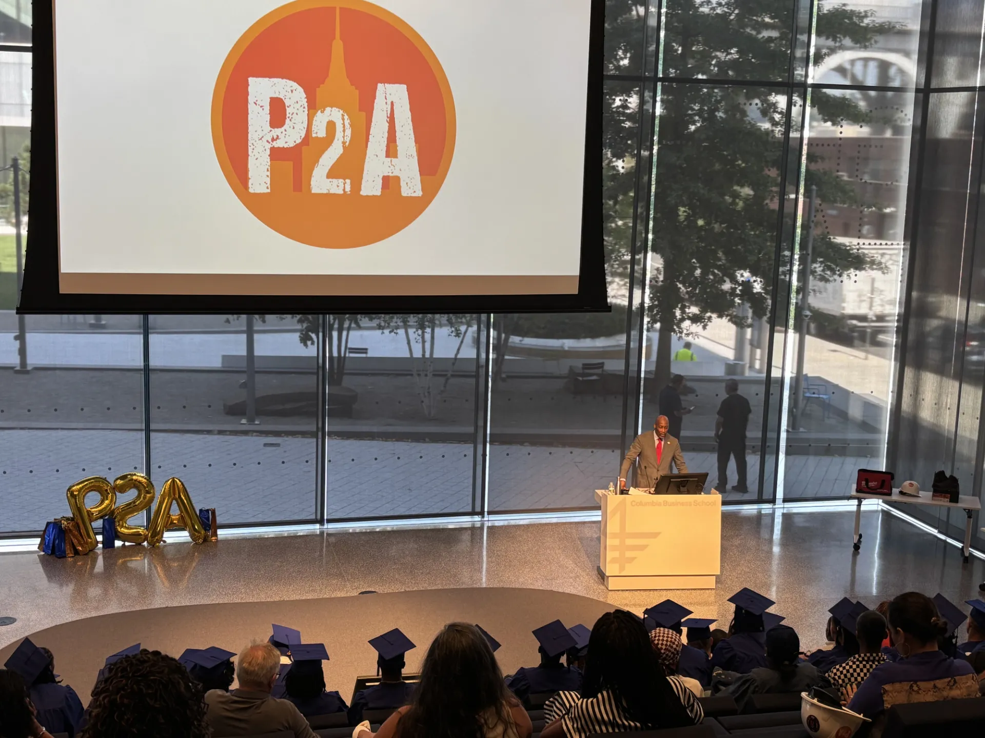 Person speaks at podium. Powerpoint at front of the room features P2A logo. Audience filled with people, some in cap and gown.