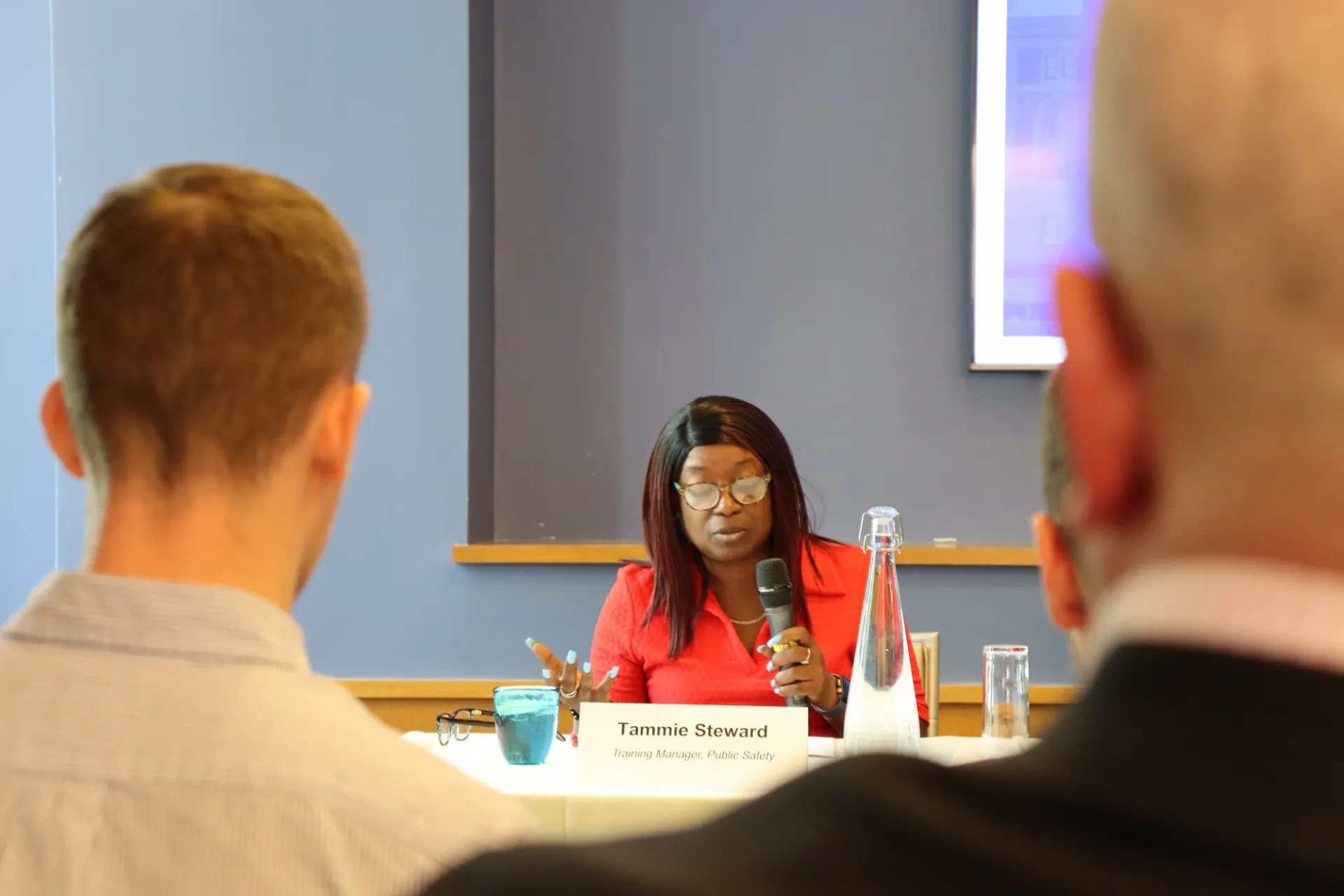 A woman speaks into a microphone at a table with a nameplate, while two people listen in the foreground.