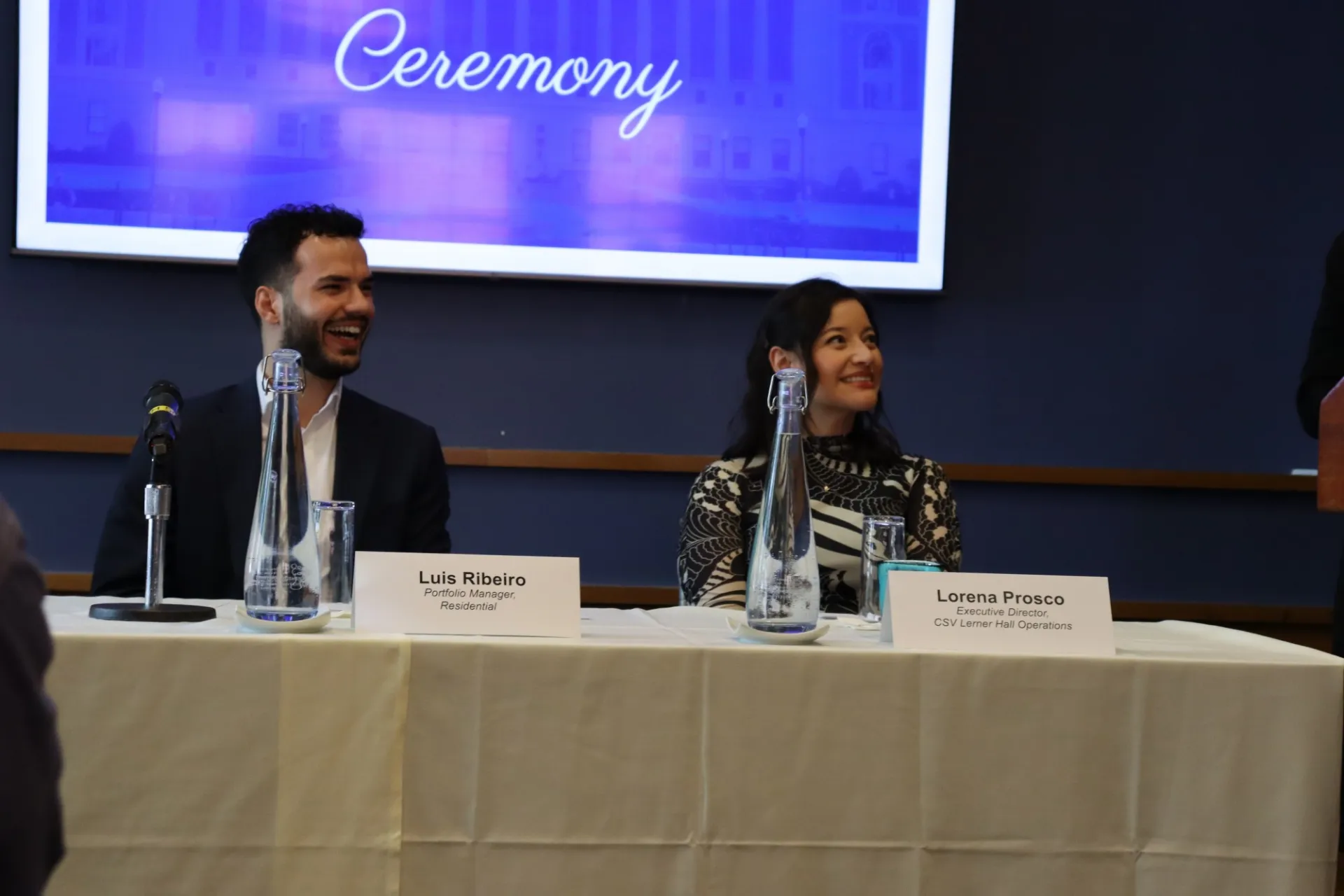 Two people sit at a table with name cards, under a screen displaying the word 'Ceremony'