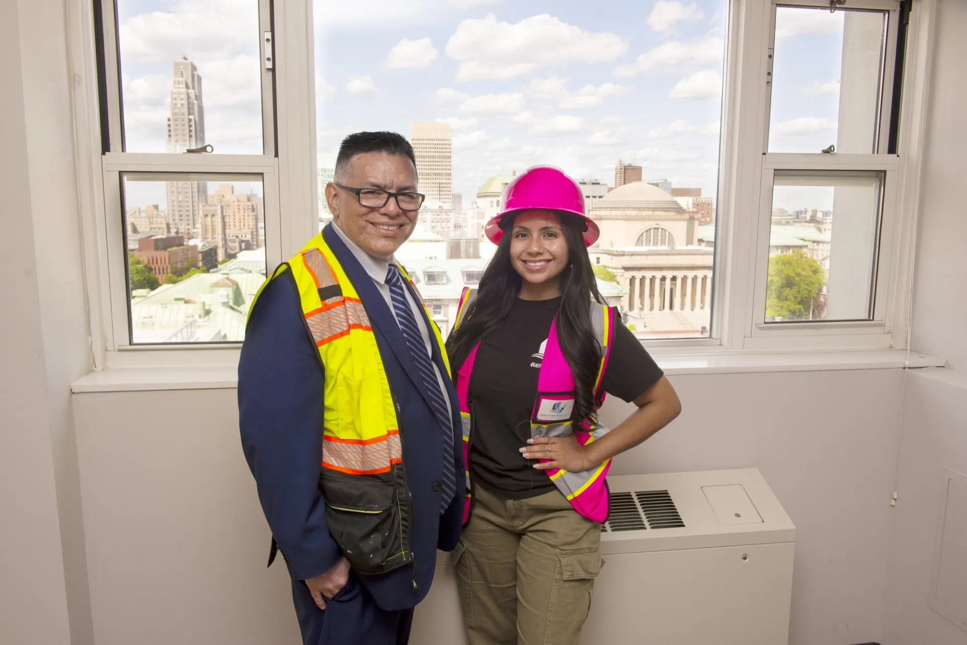 Two people in safety vests and hard hat standing in front of large windows overlooking the Morningside campus.