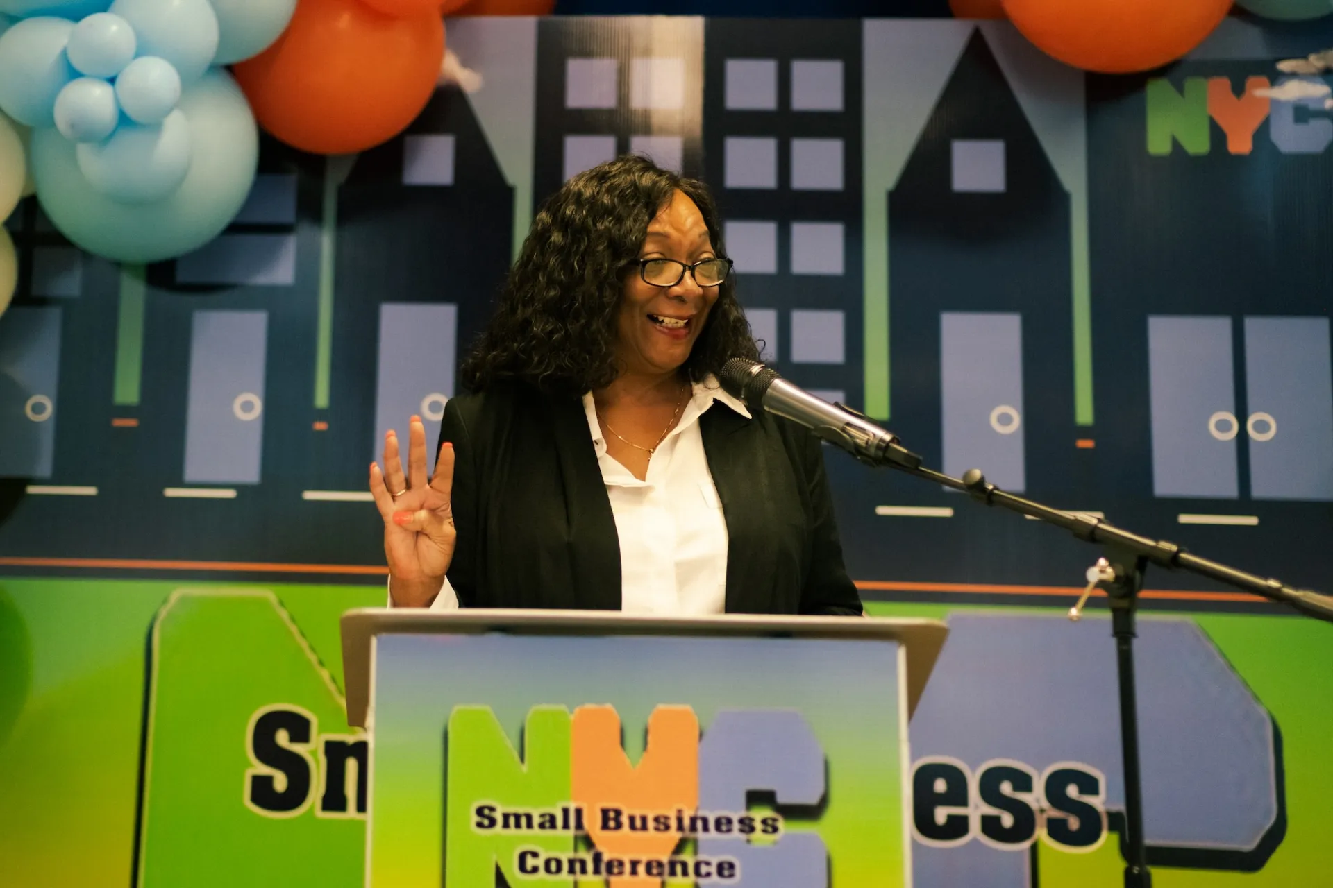 A person stands behind a podium, speaking into a microphone. The podium has a colorful sign that reads 'NYC Small Business Conference.' The background features a cityscape design with blue and orange balloons. The person is holding up four fingers on their left hand.