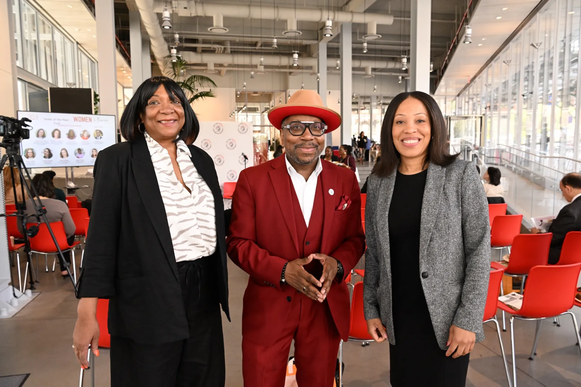 Three individuals stand together in a modern indoor space with large windows and red chairs. The person on the left wears a black blazer over a white and black patterned shirt. The person in the middle is dressed in a red suit with a matching hat and white shirt. The person on the right wears a gray blazer over a black top. In the background, people are seated and banners and posters are displayed on easels.