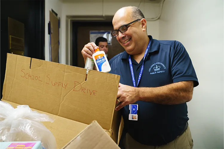 Ricardo in Public Safety shirt looking through a box labeled "school supplies" and holding Elmers Glue