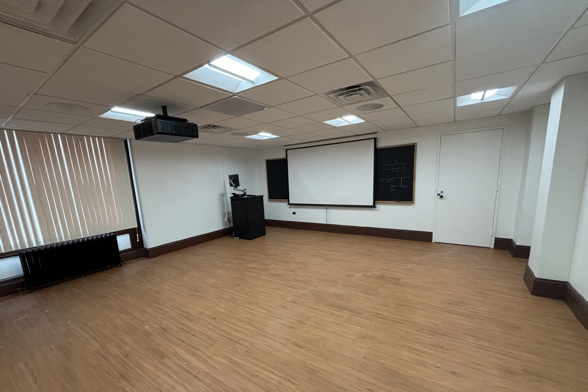 A classroom in Schermerhorn that has been refreshed with new white ceiling tiles, white paint, wooden floor planks, a new projection screen, and blackboards.