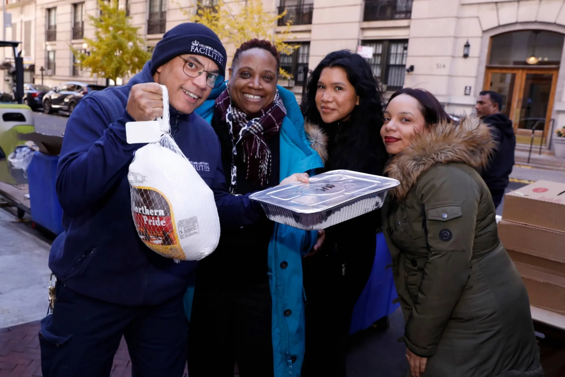 Four people stand together. One person holds a up a turkey and tray of food and wear a hat that says Facilities and Operations.
