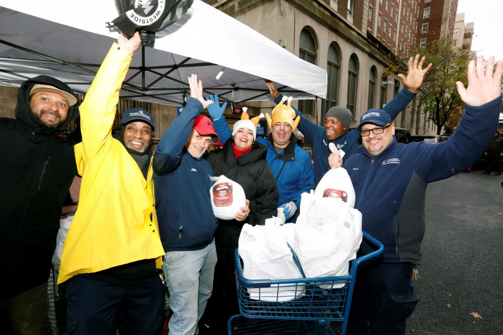 Group of people standing under a canopy tent on 114th street, raising their hands while holding turkeys and bags. One person is pushing a shopping cart filled with items. 