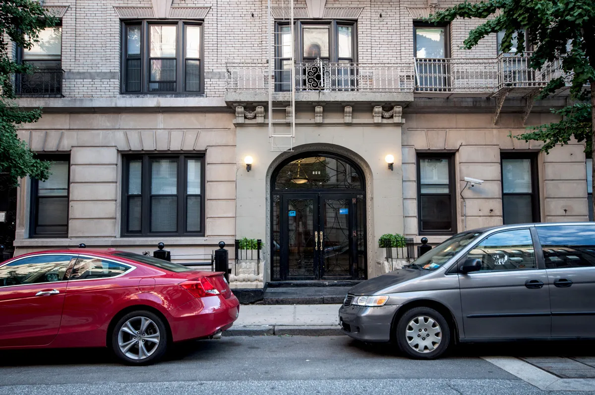 A red sedan and a grey mini van are parked on the street in front of a grey stone building.