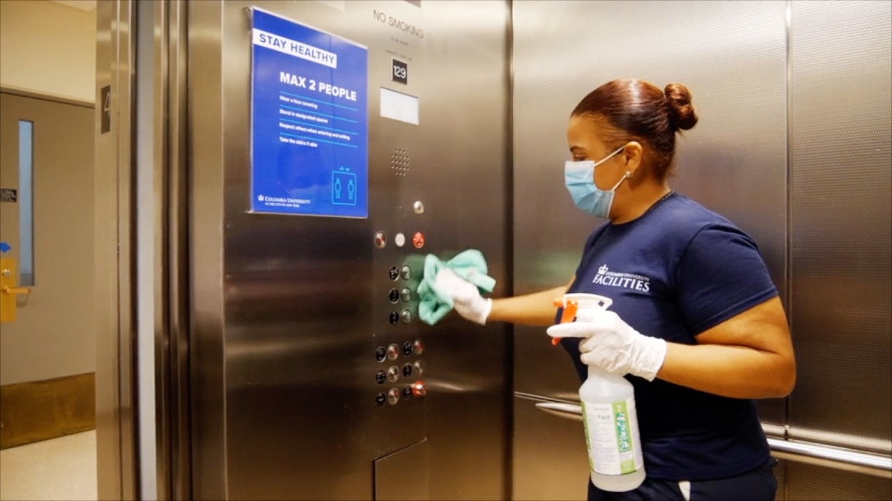 A Facilities and Operations custodial employee cleaning the inside of an elevator with a blue sign stating what the elevator occupancy is.