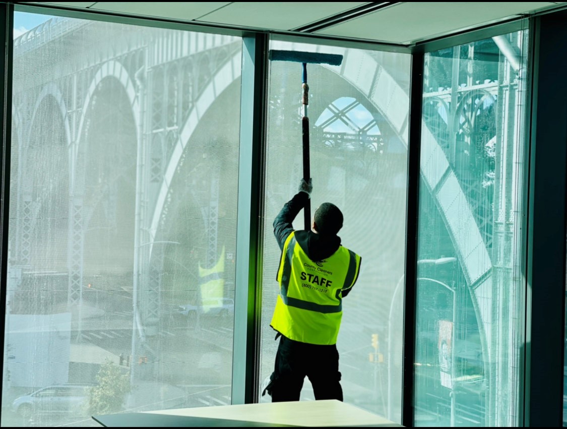 An employee of County Cleaners Corporation cleaning the inside of a tall window at Henry R. Kravis Hall using a hand-held squeegee with the Riverside Drive Viaduct visible outside the windows.