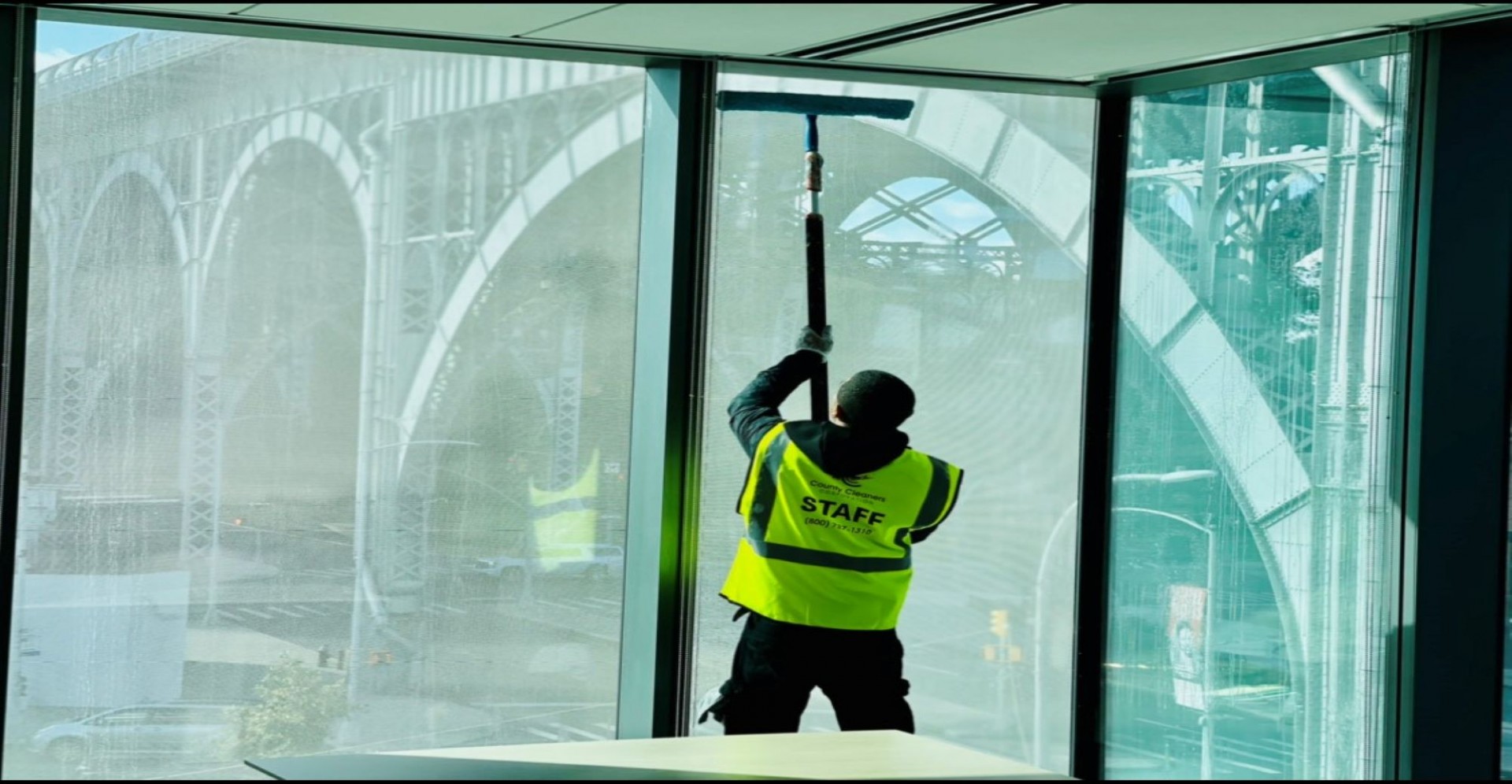 County Cleaners Corporation, an MBE participant of the ASCEND Regional Program, completing a comprehensive cleaning at the new Columbia Business School buildings. An employee of County Cleaners Corporation cleaning the inside of a tall window at Henry R. Kravis Hall using a hand-held squeegee with the Riverside Drive Viaduct visible outside the windows.