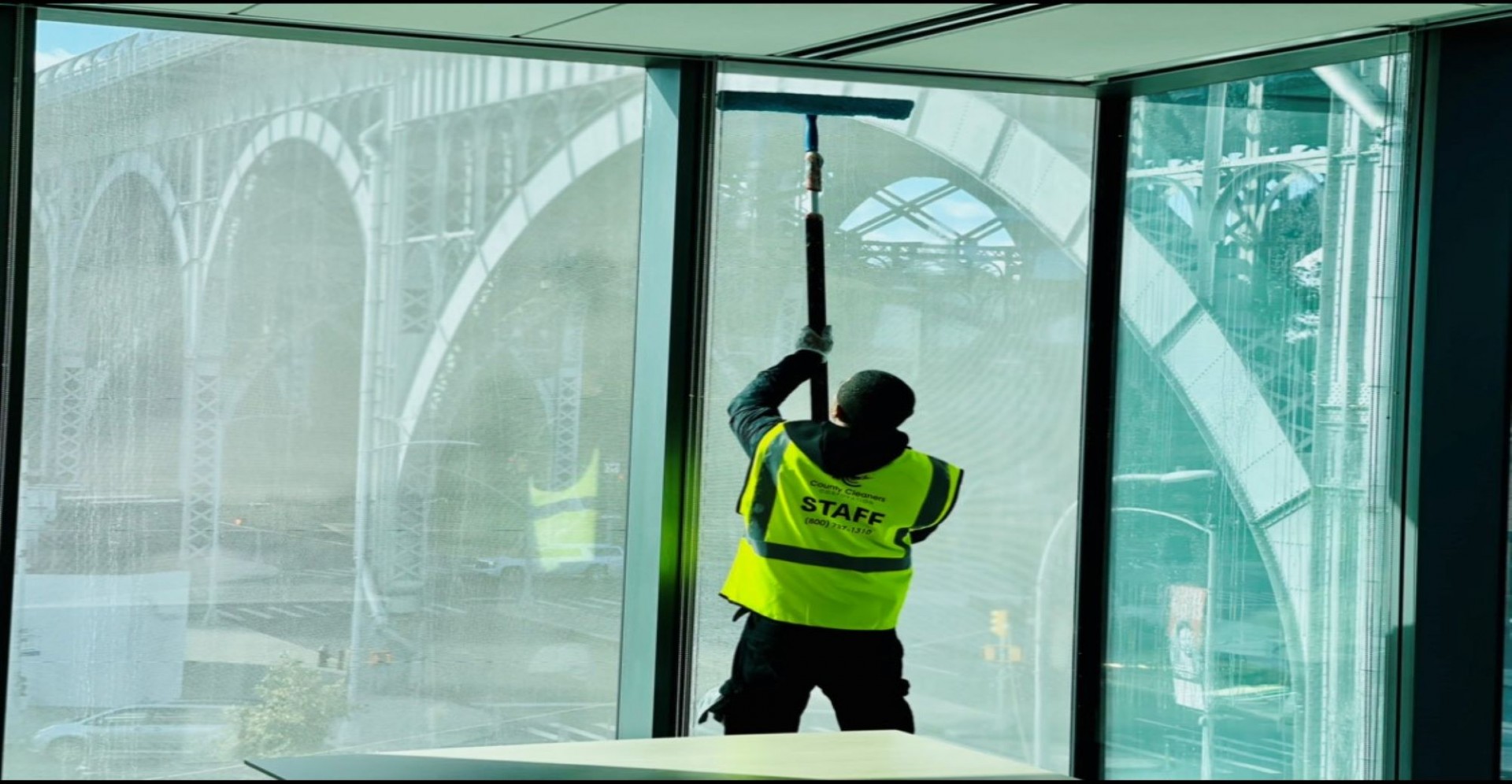 An employee of County Cleaners Corporation cleaning the inside of a tall window at Henry R. Kravis Hall using a hand-held squeegee with the Riverside Drive Viaduct visible outside the windows.