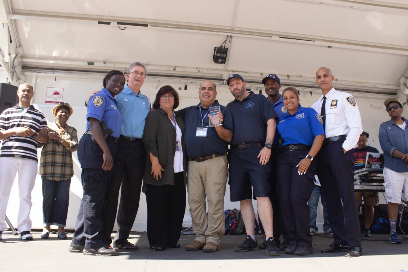 The Public Safety team gather with the NYPD on a stage, with one of the Public Safety officers holding a glass award.