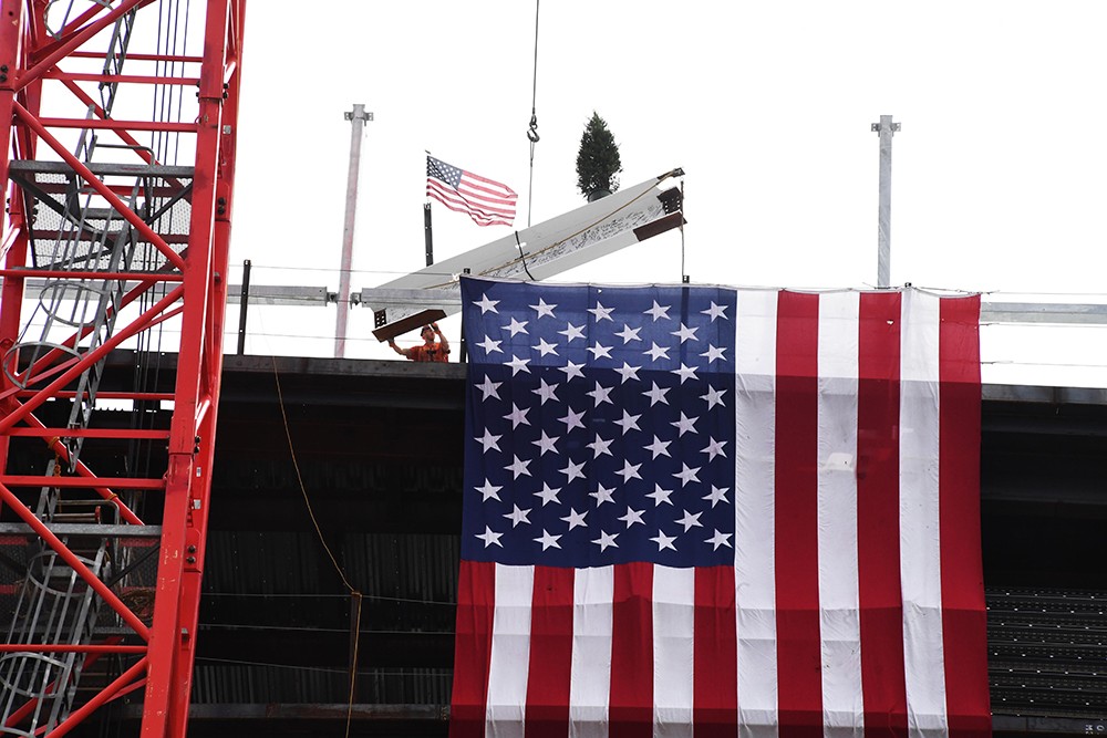 A signed beam with an American flag and fir tree being secured to the Ronald O. Perelman Center for Business Innovation.