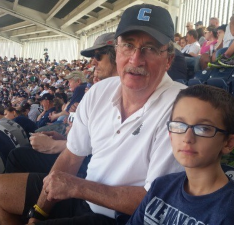 A older man wearing a white polo and baseball cap sits next to a boy in glasses. They are in an athletic stadium.