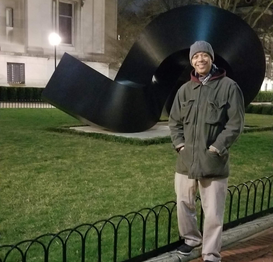 A person wearing a green jacket stands in front of an abstract sculpture.
