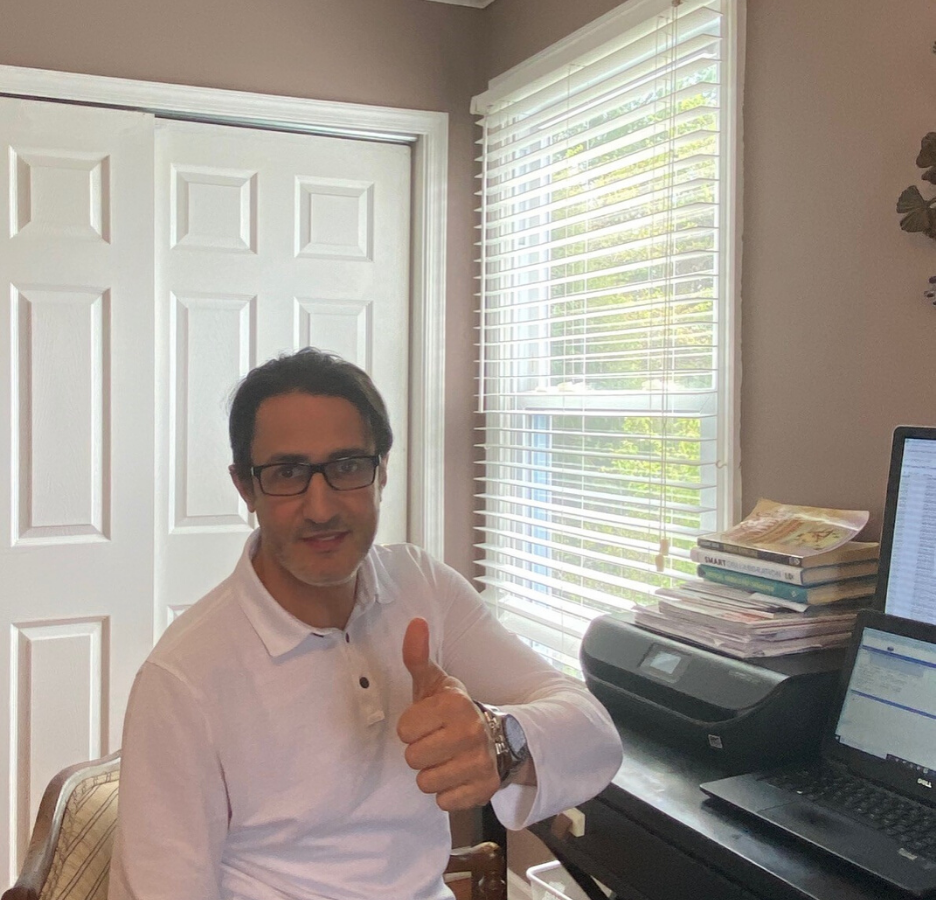 A man wearing a white button down shirt sits at a desk giving the thumbs-up sign.