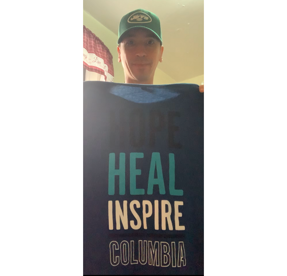 A man wearing a green baseball cap holds a shirt that says, "Hope, heal, inspire, Columbia"