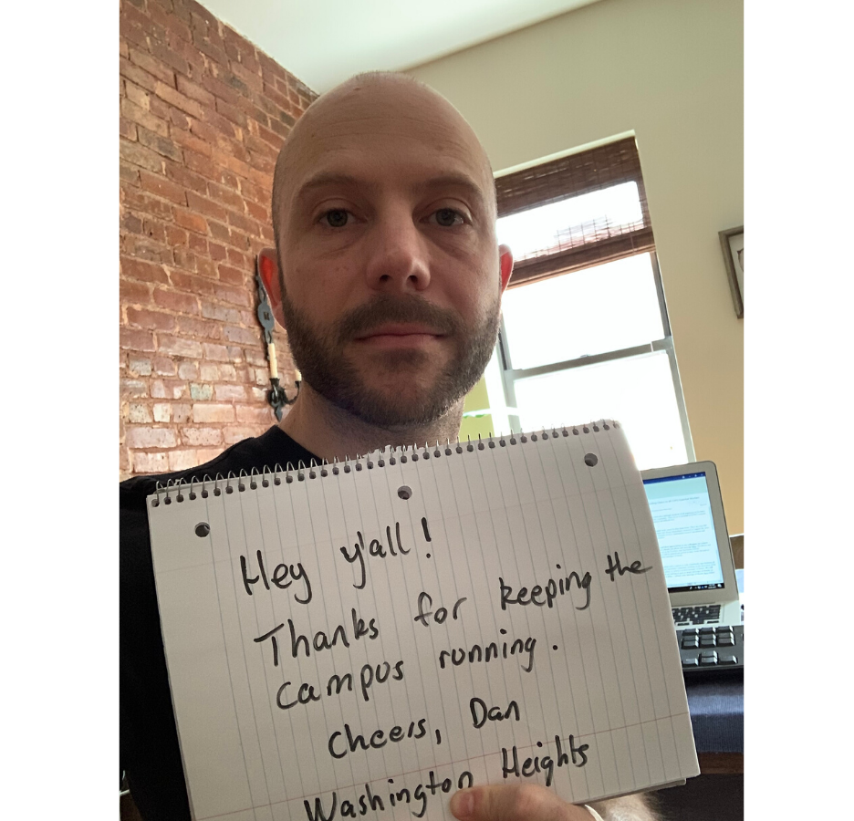 A man holding a sign that reads, "Hey y'all, thanks for keeping the campus running.  Cheers, Dan - Washington Heights" in an apartment with a brick wall