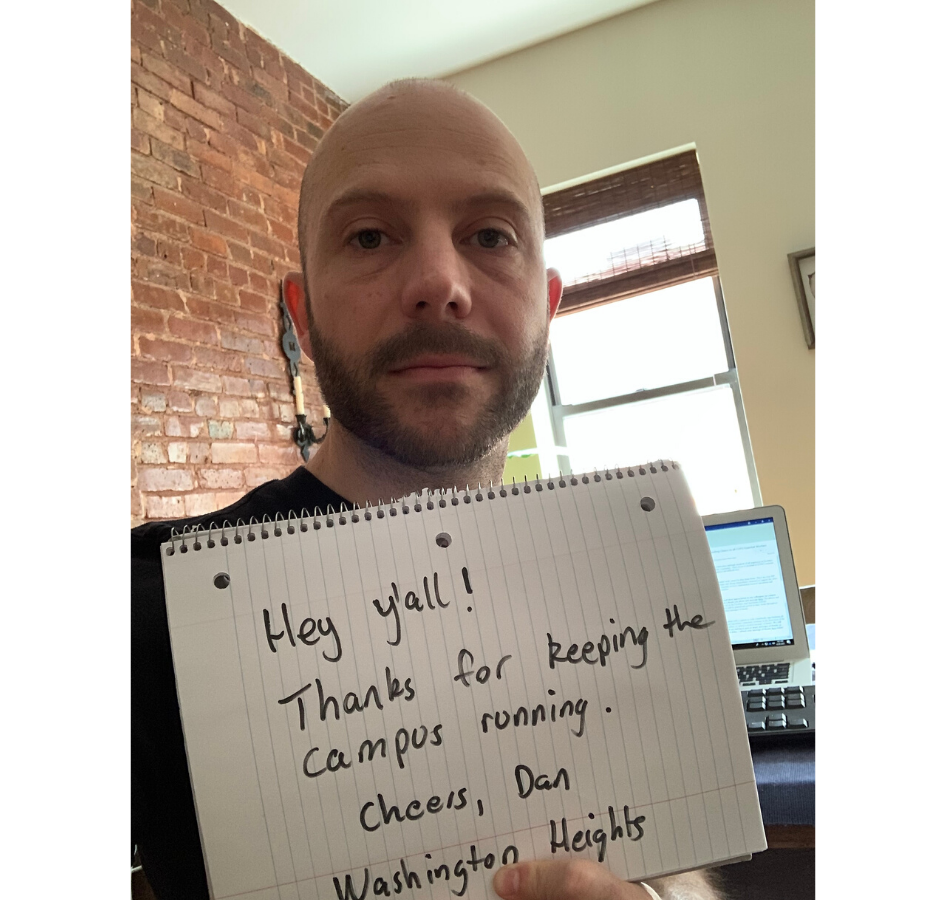 A man holding a sign that reads, "Hey y'all, thanks for keeping the campus running. Cheers, Dan - Washington Heights" in an apartment with a brick wall