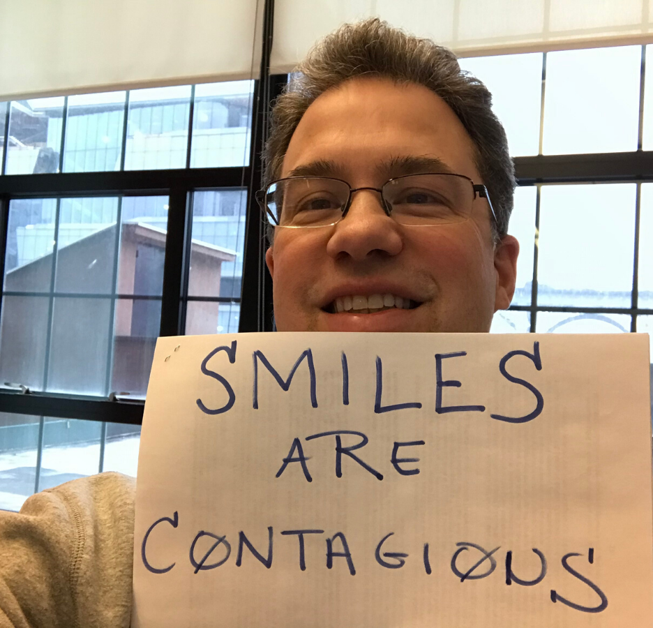 man with glasses smiles and holds up sign 