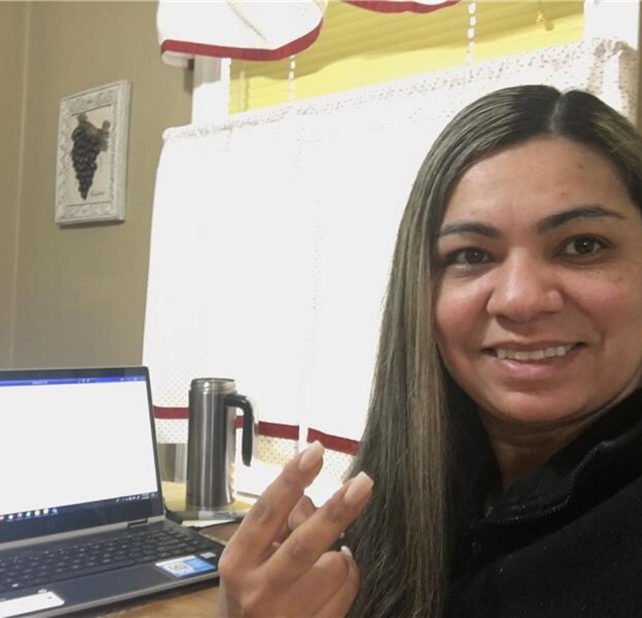 A woman makes a peace sign. She sits in front of a laptop.