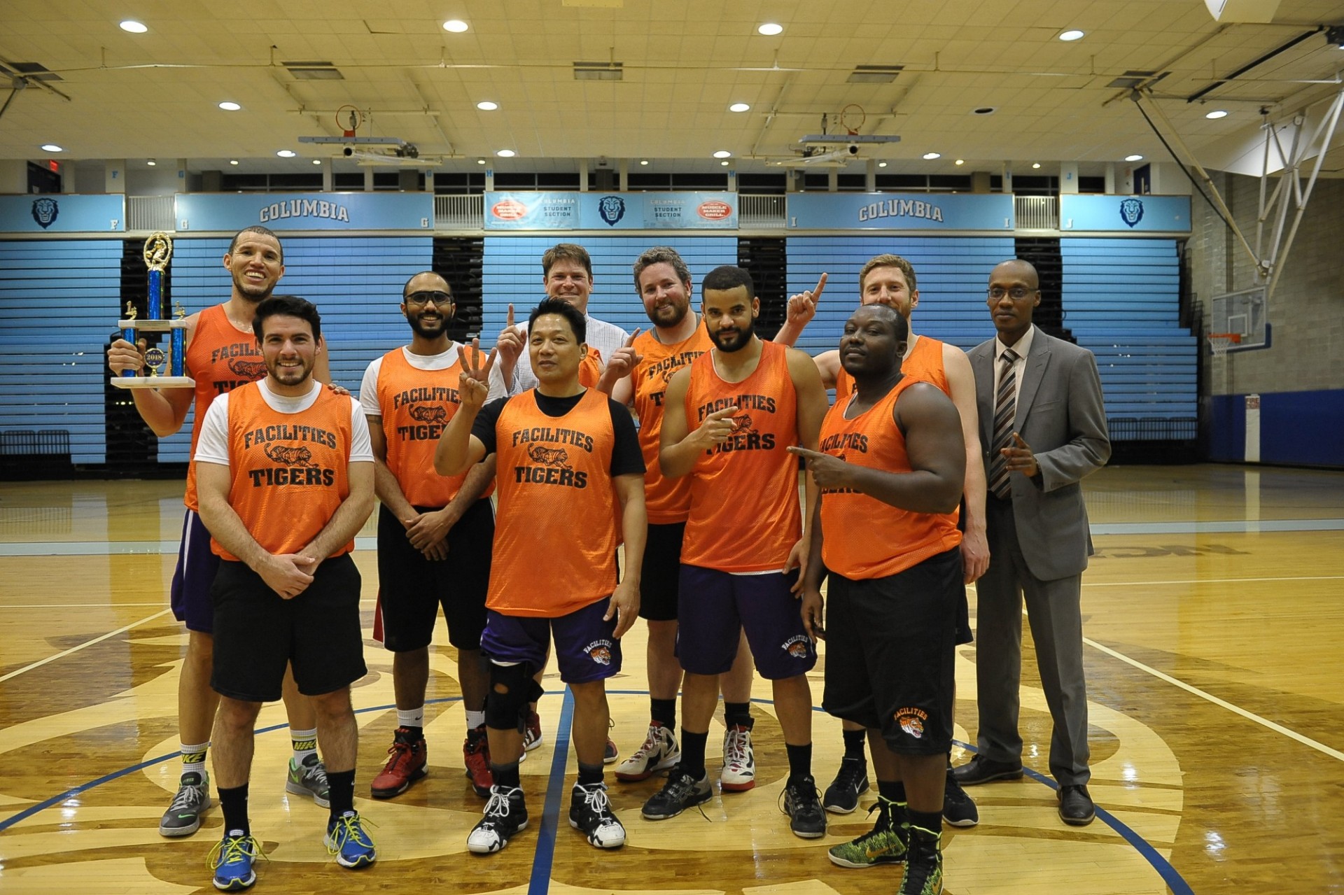 A group of people stand in orange "Facilities Tigers" jerseys inside of Levien Gym, with someone on the far left holding a trophy.