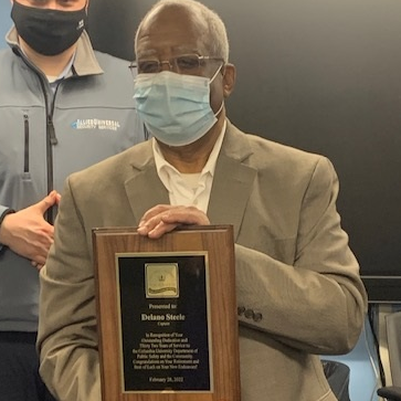 male wearing a light brown suit jacket and a face mask posing for a photo with plaque