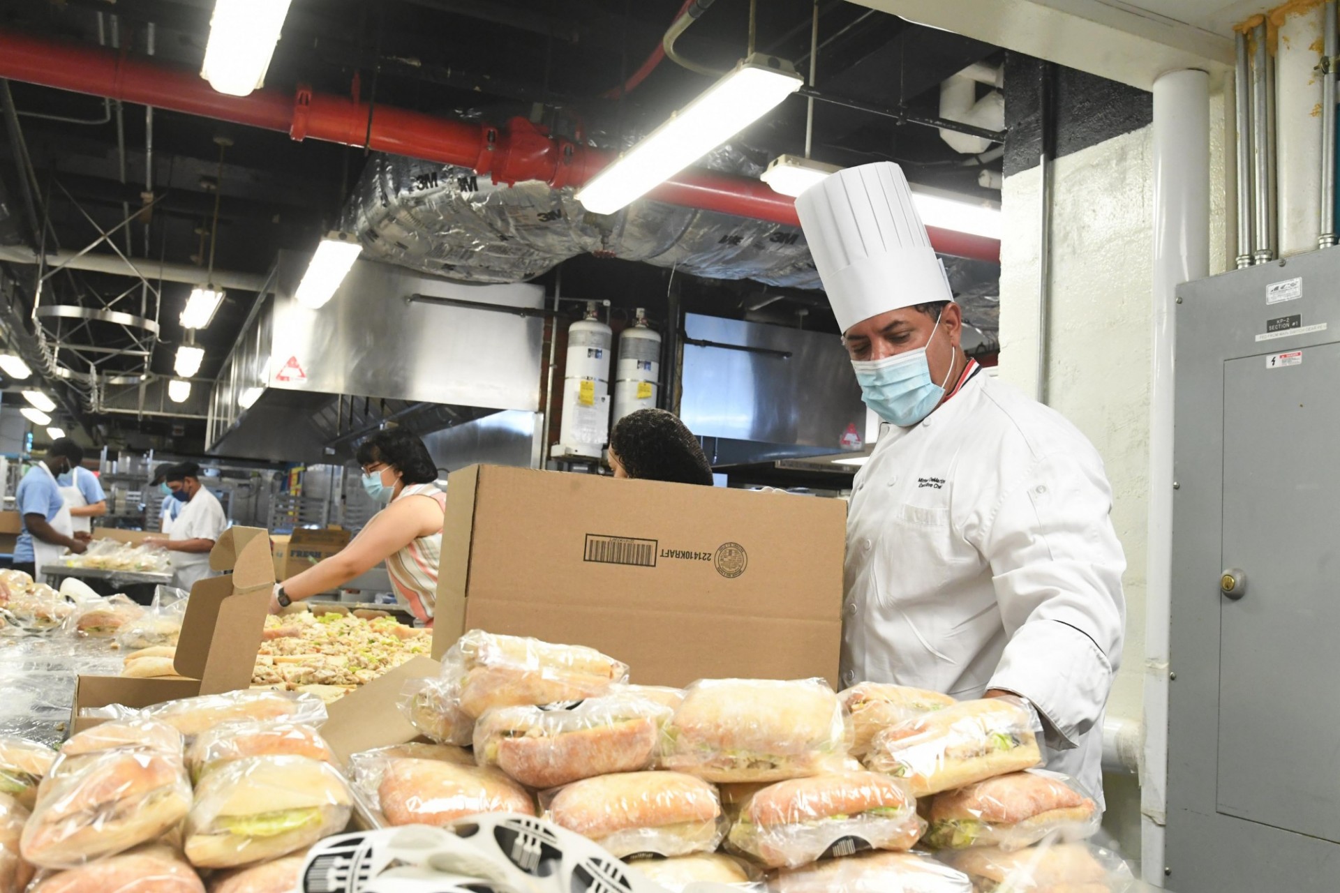 A chef is putting wrapped sandwiches in a cardboard box inside of a kitchen